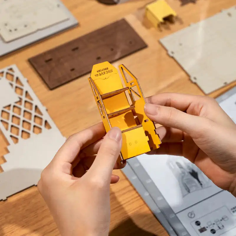 Des mains assemblent une structure miniature en bois jaune avec des étagères.