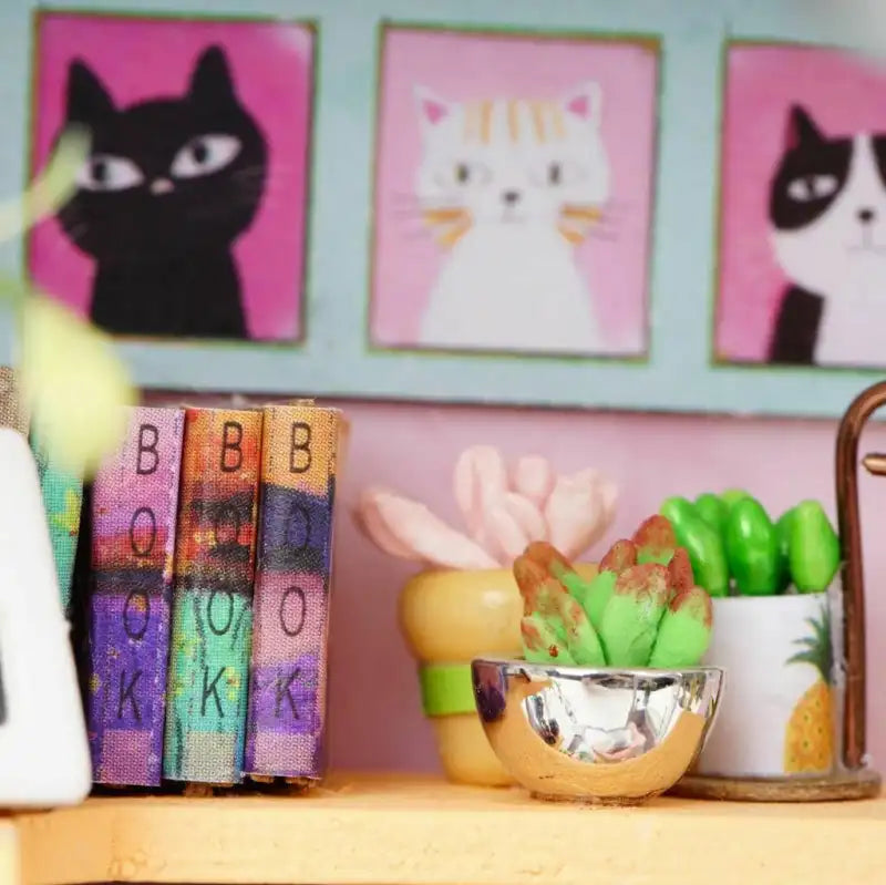 Trois portraits de chats encadrés au-dessus de livres décoratifs miniatures et de plantes grasses en pot.