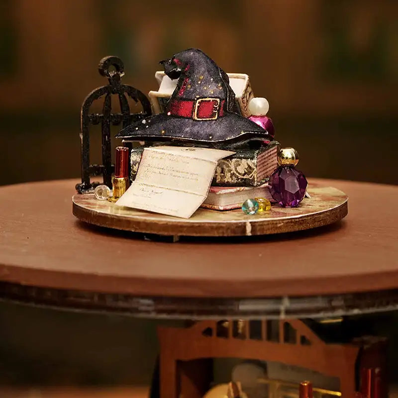 Un chapeau de sorcière miniature avec une boucle rouge, couvert de paillettes, posé sur une pile de petits livres et de papiers.