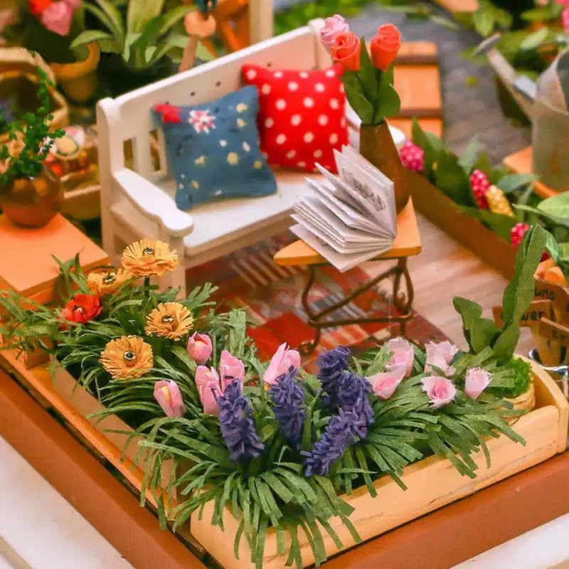Une scène de jardin miniature avec un banc blanc, des fleurs en pot et un livre ouvert sur une petite table.