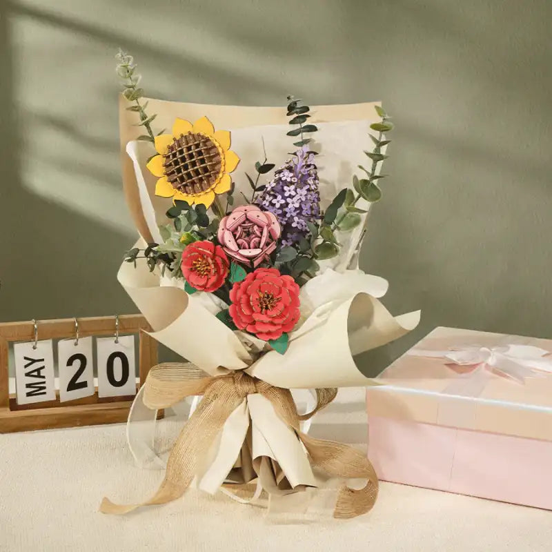 Un bouquet de fleurs artificielles de couleurs et de styles différents, enveloppé dans du papier et noué avec un ruban en toile de jute, accompagné d’une boîte cadeau rose.