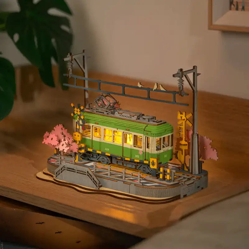 Un train miniature en bois vert illuminé avec des cerisiers en fleurs et des poteaux électriques.