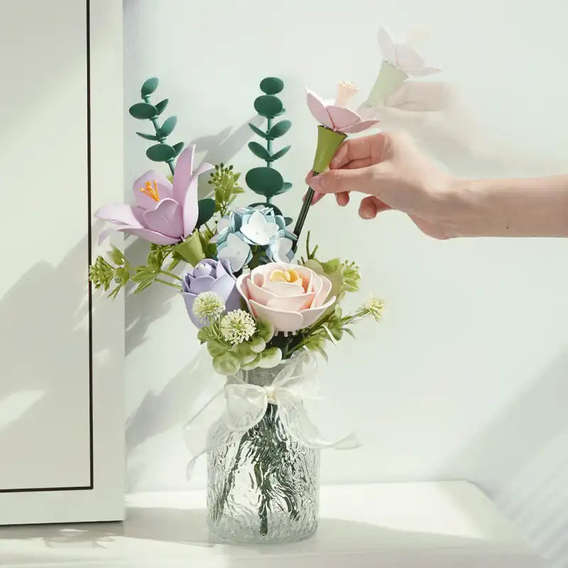 Un bouquet de fleurs artificielles dans des tons pastel, comprenant des roses, des crocus et des feuilles d’eucalyptus, disposées dans un vase en verre texturé transparent avec un ruban blanc.