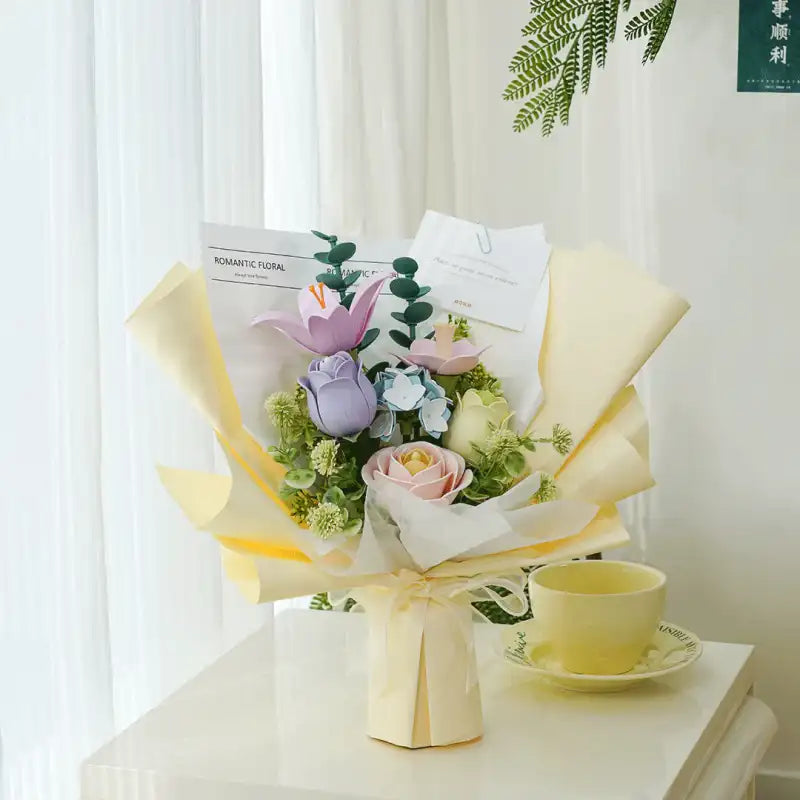 Un bouquet de fleurs artificielles de couleur pastel avec du feuillage vert, enveloppé dans du papier jaune clair et blanc, noué avec un ruban.