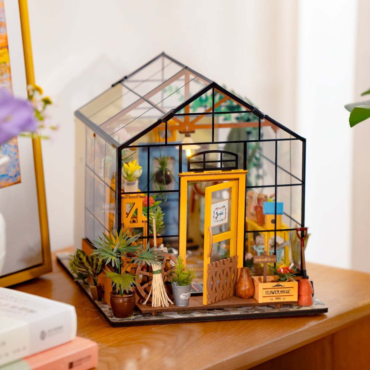 Une serre miniature en bois avec des parois transparentes et une porte jaune ouverte, remplie de petites plantes et d’outils de jardinage.