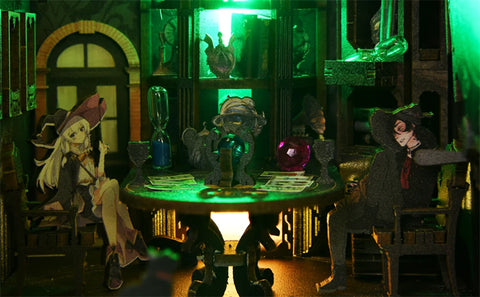 Diorama représentant deux sorcières de style animé assises à une table dans une pièce éclairée en vert et dotée de divers objets magiques.