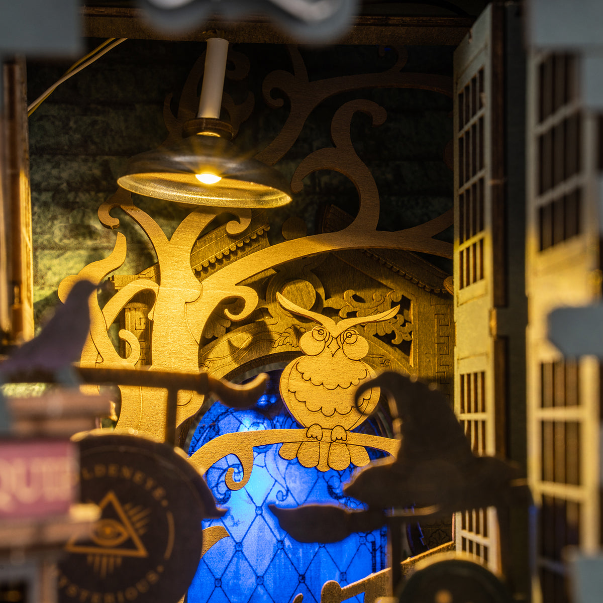 Figurine de hibou en bois sculpté sur une branche, éclairée par une lampe suspendue, avec une fenêtre à motifs bleus derrière elle.